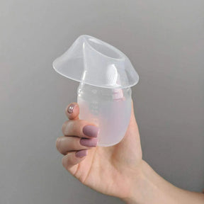 Hand holding a clear silicone breast pump for milk collection against a plain background.