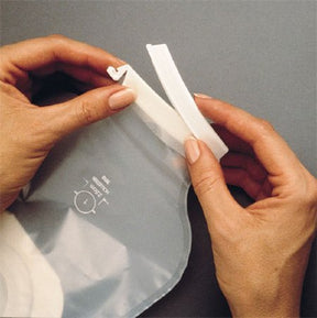 Hands attaching a plastic clamp to a transparent ostomy pouch on a gray background.
