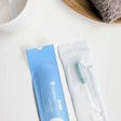 Toothbrush in sealed packaging on white surface, next to cup and towel, medical supply delivery
